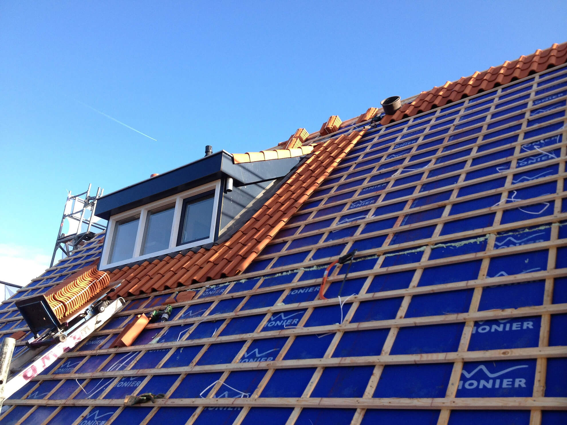 Nieuwe oranje dakpannen worden gelegd op panlatten en blauwe onderdakfolie, met een dakkapel